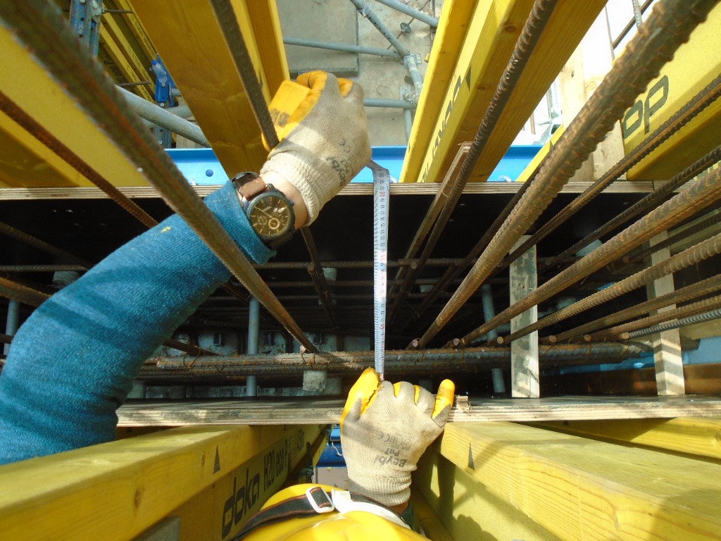 A construction professional using a tape measure to take precise measurements within a wooden formwork structure reinforced with steel rebar.